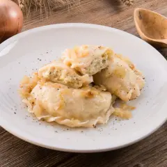 Pierogi ruskie na białym talerzu, posypane pieprzem, obok cebula i drewniane sztućce.