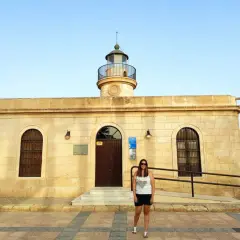 Mujer sonriente posa frente al Faro de Roquetas de Mar, con el mar azul de fondo.