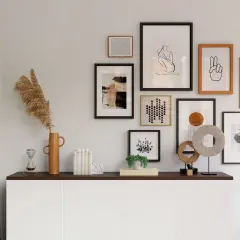 Galeria obrazów nad komodą w salonie, w tym abstrakcje, grafiki i sztuka liniowa. Wśród dekoracji wazon z trawami, klepsydra i wisząca roślina.