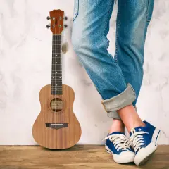 Drewniane ukulele stoi obok osoby w jeansach i niebieskich trampkach. To małe, czterostrunowe ukulele.
