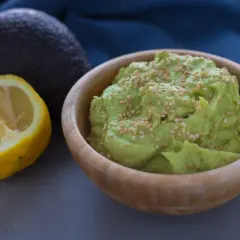 Hummus di avocado cremoso in ciotola di legno, guarnito con semi di sesamo. Accanto, un avocado intero e mezzo limone.