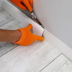 Mani con guanti arancioni che usano un taglierino per rifinire un battiscopa bianco, pronto per incollare.