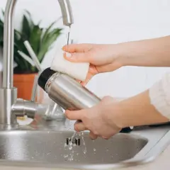 Hände reinigen eine Edelstahl-Thermoskanne unter fließendem Wasser im Spülbecken.