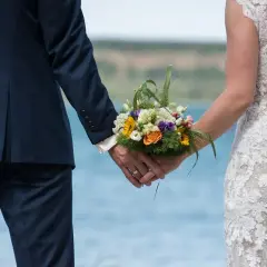 Para młoda trzyma się za ręce, trzymając bukiet kwiatów. Ceremonia ślubna w plenerze.