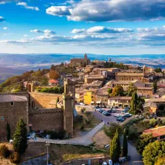 A panoramic view of medieval towns in Italy, perched on a hilltop under a bright blue sky with fluffy clouds.