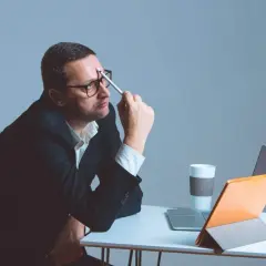 Mężczyzna w okularach siedzi przy biurku, zamyślony, trzymając długopis przy skroni. Obok laptop, tablet i kubek. Zastanawia się nad ryczałtem.
