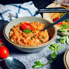 Schüssel mit aromatischem griechischem Reis, garniert mit Tomaten und Petersilie. Frische Tomaten und Knoblauch im Hintergrund.