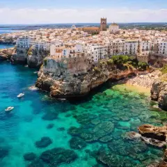 Vue aérienne de Polignano a Mare, avec ses maisons blanches perchées sur des falaises rocheuses surplombant une mer turquoise et une plage bondée.