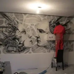 Artysta w czerwonym kombinezonie tworzy malarstwo artystyczne wnętrz – surrealistyczny mural z twarzami i mechanicznymi elementami.