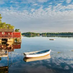 En vit båt ligger förtöjd vid en röd sjöbod. Kanske är det en båt till Drottningholm, redo för en lugn tur på vattnet.