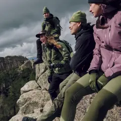 Grupa przyjaciół w zielonych i fioletowych kurtkach softshell siedzi na skale, podziwiając górski krajobraz.