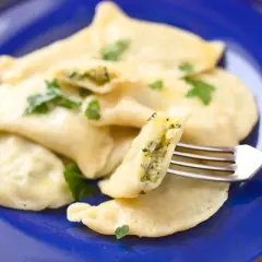 Pyszne pierogi ze szpinakiem, serem i czosnkiem, udekorowane natką pietruszki, na granatowym talerzu. Jedna porcja jest przekrojona widelcem.