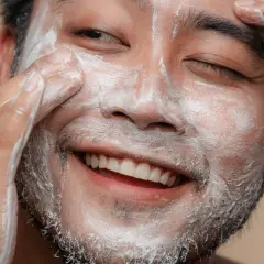 Un visage d'homme souriant, couvert de mousse nettoyante, se fait un soin du visage.