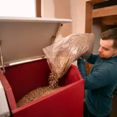 Mężczyzna wsypuje pellet do zasobnika kotła. Czy to tańsze niż gaz? W tle widać worki z opałem.