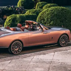Elegancki Rolls-Royce Boat Tail w kolorze miedzianego różu, z drewnianym tyłem, zaparkowany wśród bujnej zieleni.