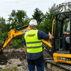 Instruktor pokazuje, jak szybko można ukończyć kurs na koparkę. Operator w kamizelce "Akademia Operatora" uczy się obsługi maszyny JCB.