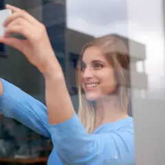 Uśmiechnięta blondynka robi sobie selfie w lustrze, trzymając telefon. Wskazówka, jak zrobić dobre selfie w lustrze: złap światło!