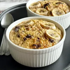 Dwie porcje owsianki z bananem i kawałkami czekolady w białych kokilkach na czarnej tacy. Idealne na słodkie śniadanie.