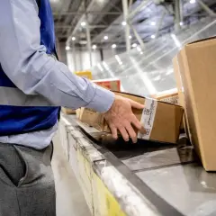Pracownik magazynu w kamizelce odblaskowej układa paczki na taśmie. Na jednej z nich widoczny jest kod kreskowy ean.
