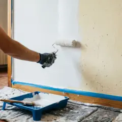 Persona con guantes pinta pared de blanco con rodillo. Bandeja con pintura y brocha lista para seguir como pintar una pared.
