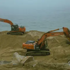 Dwie koparki pracują na piaszczystym brzegu. Widoczny jest element roboczy koparki, gotowy do pracy.