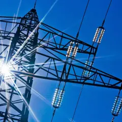 Słońce przebija się przez kratownicę wieży energetycznej, oświetlając izolatory przewodów, które przenoszą prąd dwufazowy.