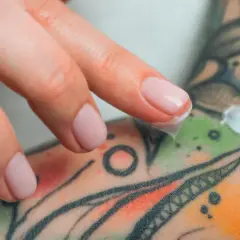 Main d'une personne appliquant une crème sur un tatouage coloré. Si vous suspectez une infection tatouage, que faire ? Consulter un professionnel de santé.