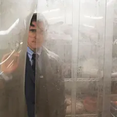 Un uomo in giacca e cravatta, con occhiali, spia da dietro una tenda di plastica. Potrebbe essere una scena da "La casa di Jack", spiegazione inquietante.