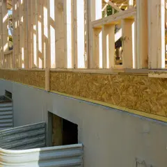 Budowa domu w stanie surowym, widoczny betonowy fundament, okna piwniczne i drewniana konstrukcja ścian. Brak cokołu na elewacji.