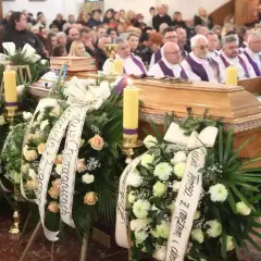 Ksiądz odczytuje ostatnie pożegnanie zmarłego przy trumnie, otoczonej wieńcami i świecami.