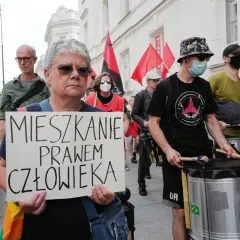 Kobieta trzyma transparent: "MIESZKANIE PRAWEM CZŁOWIEKA". W tle protestujący z bębnami i flagami.