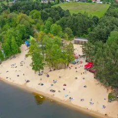 Plaża nad zalewem Cedzyna tętni życiem. Ludzie odpoczywają na piasku, ciesząc się słońcem i widokiem na jezioro.