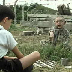 Chłopiec w pasiastej koszuli wskazuje na planszę do gry. Obok siedzi inny chłopiec w białej koszuli i szelkach.
