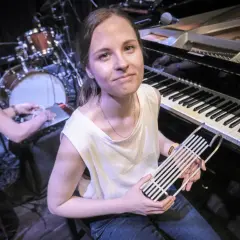 Kobieta z instrumentem muzycznym przed fortepianem i perkusją.