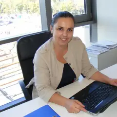 Uśmiechnięta kobieta w beżowej marynarce siedzi przy biurku z laptopem.