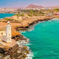 Faro blanco en una costa rocosa con aguas turquesas. Un pueblo costero se extiende detrás. ¡Ideas de qué hacer en Cabo Verde!