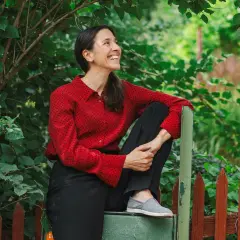 Paloma Grandin sitter avslappnat vid ett staket, omgiven av grönska. Hon ler och tittar uppåt, fångad i ett ögonblick av glädje.