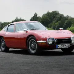 Klasyczny, czerwony Toyota 2000GT, jeden z najbardziej pożądanych japońskich samochodów sportowych, prezentuje swoje eleganckie linie na tle zieleni.