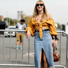 Mujer sonriente con un outfit falda vaquera larga, blusa amarilla anudada y bolso marrón.