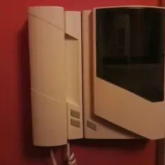 Biały unifon zamontowany na czerwonej ścianie. Urządzenie ma prosty, nowoczesny design z ciemną szybką.