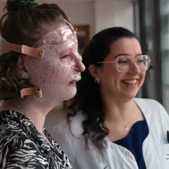 Une femme porte un masque transparent sur le visage, peut-être pour aider à comment cacher une brûlure. Une autre femme, souriante, la regarde.