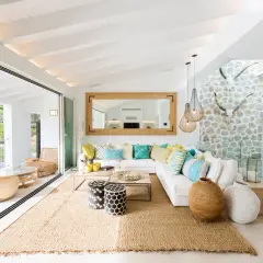 Helles Wohnzimmer mit Finca Style Einrichtung, weißem Sofa, bunten Kissen, Jute-Teppich und Blick auf das Meer.