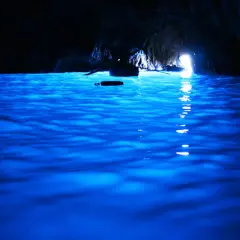 Acqua blu elettrico nella grotta, un piccolo remo galleggia mentre una barca si dirige verso la luce all'uscita della blaue grotte.