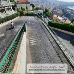 Le 13 discese di Napoli, una vista panoramica di una strada in discesa con un motorino, tra palazzi e vegetazione, che porta verso il mare.