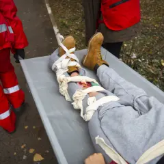 Na noszach leży osoba z opatrzonym złamaniem otwartym nogi. Ratownicy w czerwonych strojach udzielają pomocy.