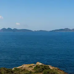 El mar azul se extiende hasta las islas de **cabo Estai**, con una costa rocosa y vegetación en primer plano.