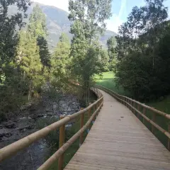 Paseo de madera junto a un río, parte de la ruta del ferro andorra, rodeado de árboles y montañas.