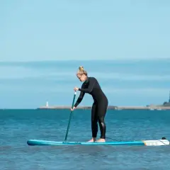 Kobieta w piance pływa na desce SUP po spokojnym morzu. W tle widać latarnię morską i wybrzeże.