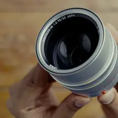 Hands hold a Leica Noctilux M 50 mm lens, its silver barrel and prominent f/0.95 aperture gleaming.