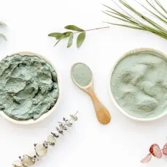 Dos recipientes con arcilla verde, uno en polvo y otro en pasta, junto a una cuchara de madera y ramas secas. Descubre las arcilla verde propiedades.